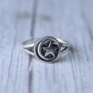 May include: A silver ring featuring a crescent moon and a five-pointed star within a circular frame. The ring has a split band design. The ring is likely made of sterling silver or a similar metal.