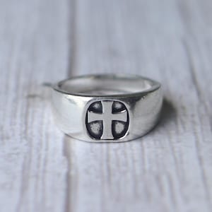 Puede incluir: Anillo plateado con diseño de cruz. El anillo presenta una banda redondeada y una cara rectangular con un emblema de cruz en relieve. La cruz está centrada, creando un aspecto clásico. El anillo es probablemente de metal.
