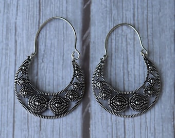 May include: A pair of silver-toned hoop earrings. Each earring features a crescent-shaped design with intricate openwork and circular floral patterns. The earrings have a hook closure and are displayed against a light wood-grain background.