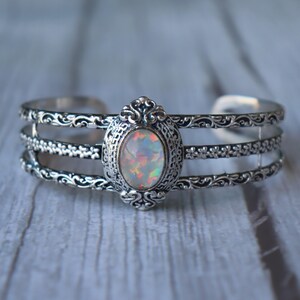 Peut inclure: Bracelet manchette en argent orné d'une pierre d'opale ovale. Le bracelet présente un design orné avec trois bandes parallèles et des volutes décoratives. L'opale affiche un jeu de couleurs, dont le blanc, le rose et le vert.