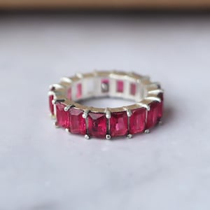 May include: A silver ring with a continuous row of vivid, rectangular, ruby-red gemstones. The gemstones are set closely together, creating a striking design. The ring is a statement piece, ideal for adding a splash of colour to any outfit.