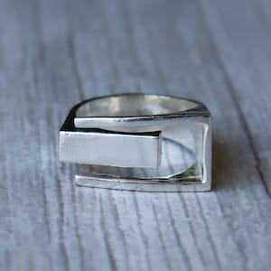 Könnte beinhalten: Ein silberfarbener Ring mit einem geometrischen Design. Der Ring hat einen rechteckigen Balken, der sich von dem quadratischen Band erstreckt. Der Ring besteht aus einem glänzenden, metallischen Material.