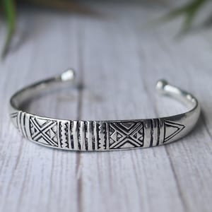 May include: A silver cuff bracelet with etched geometric patterns. The open-ended bracelet features repeating shapes, including triangles, X's, and vertical lines. The bracelet is displayed on a light-coloured wooden surface.