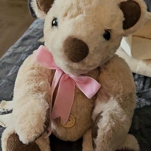 May include: A plush teddy bear with light beige fur, featuring dark brown accents on the ears, nose, and paw pads. The bear has black plastic eyes and a light pink satin ribbon tied in a bow around its neck, with a small wooden button on its chest.