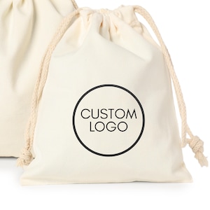 Puede incluir: Bolsa con cordón de color crema con un logotipo circular negro que dice "CUSTOM LOGO". La bolsa está hecha de una tela suave y texturizada y tiene un cierre de cordón de color natural. Otra bolsa es parcialmente visible en el fondo.