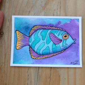 Pode incluir: Uma pintura em aquarela de um peixe colorido com corpo turquesa e detalhes em azul e roxo. O peixe tem uma cauda amarela e está sobre um fundo azul e roxo. A obra está assinada.