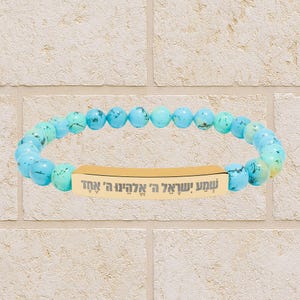 Könnte beinhalten: Ein Armband mit türkisfarbenen Perlen und einem goldfarbenen rechteckigen Mittelstück. Das Mittelstück trägt hebräischen Text. Das Armband wird vor einer hellfarbigen Backsteinmauer präsentiert.