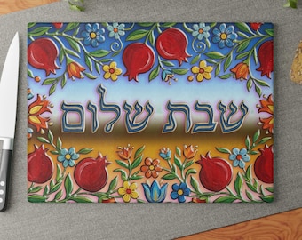 May include: Rectangular cutting board with a colorful floral and pomegranate design. The board features the Hebrew text "Shabbat Shalom" in the center, surrounded by red pomegranates, blue, yellow, and orange flowers, and green leaves.