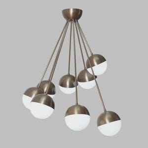 Può includere: Un lampadario moderno con finitura bronzo. L'apparecchio presenta più luci sferiche, ciascuna con una metà inferiore bianca e una metà superiore bronzo. Le luci sono sospese da sottili aste di bronzo.