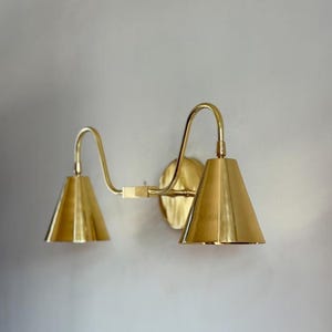 Peut inclure: Applique murale en laiton avec deux abat-jours coniques. Le luminaire présente un design incurvé avec une plaque de montage circulaire. Les abat-jours sont de couleur or chaud et le luminaire est monté sur un mur de couleur neutre.