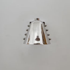 Peut inclure: Applique murale argentée polie avec un abat-jour en forme de cloche. L'applique comporte de petits éléments décoratifs ronds sur les côtés. Le design est moderne et minimaliste, adapté à l'éclairage intérieur.