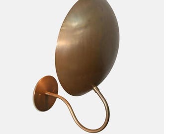 May include: A bronze-colored wall sconce with a large, oval shade and a curved arm. The sconce has a circular base and a warm, metallic finish. The design is minimalist and modern, suitable for interior lighting.