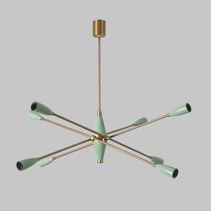 Może przedstawiać: Nowoczesny żyrandol w stylu mid-century, z mosiężnym wykończeniem i miętowo-zielonymi akcentami. Oprawa oświetleniowa ma wiele ramion wychodzących ze środka, każde zakończone stożkowym światłem. Całość jest symetryczna i elegancka.