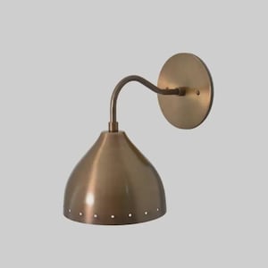 May include: A bronze-colored wall sconce with a curved arm and a conical shade. The shade has a series of small, evenly spaced holes near the bottom. The sconce is mounted on a round backplate. This fixture is suitable for interior lighting.