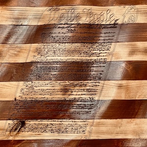 May include: Wooden American flag with the text "We the People" and the Constitution etched into the wood. The flag features alternating stripes of light and dark brown, with a blue section in the upper left corner.