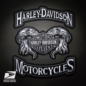 Peut inclure: Trois écussons Harley-Davidson noirs et blancs. Le patch supérieur indique "HARLEY-DAVIDSON" dans une bannière incurvée. Le patch central présente deux têtes d'aigle flanquant un logo qui dit "MOTOR HARLEY-DAVIDSON CYCLES". Le patch inférieur indique "MOTORCYCLES".