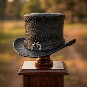 May include: A black top hat featuring a textured snakeskin pattern across the crown and brim. A smooth black leather band encircles the base, accented with brown braided leather details and a central silver-toned emblem of two crossed revolvers.