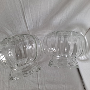 May include: Two clear glass lamp shades featuring a rounded, bulbous shape with vertical ribbed detailing and a ruffled, scalloped edge at the base. These transparent glass fixtures have a wide opening at the top and a smaller opening at the bottom.