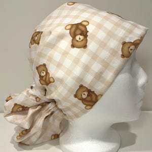 May include: A beige and white checkered scrub cap featuring a repeating pattern of brown teddy bears. The cap is tied in the back and is designed to cover the hair. The fabric has a soft, slightly textured appearance.