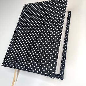 May include: A black and white polka dot journal with a cream-colored ribbon bookmark. The journal is open, revealing the white pages inside. The cover is a dark black with white polka dots.