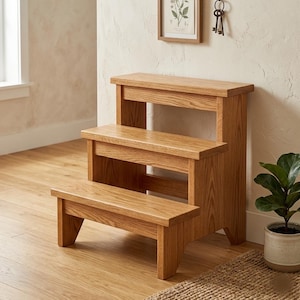 May include: A three-step wooden step stool with a natural light brown wood grain finish. The stool features a wide base, two lower steps, and a higher top platform, all constructed with sturdy vertical side supports and horizontal treads.