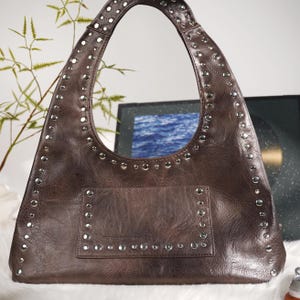 May include: Brown shoulder bag with silver-colored studs. The bag has a large, curved handle and a front pocket. The studs are evenly spaced along the edges of the bag and pocket, adding a decorative element. The bag appears to be made of a textured material.
