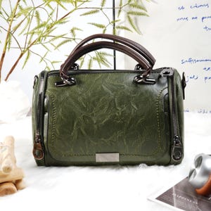 Może przedstawiać: Zielona skórzana torebka z teksturowanym wykończeniem. Torba ma górny uchwyt z brązowymi akcentami, prostokątny kształt i srebrną metalową płytkę z przodu. Posiada dwa boczne zamki błyskawiczne.