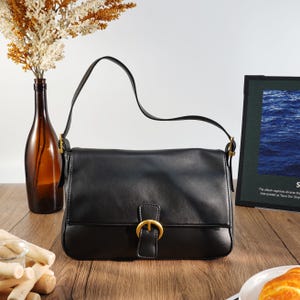 Pode incluir: Bolsa de ombro preta com fecho de fivela dourada. A bolsa tem uma alça longa e formato retangular. Uma garrafa de vidro marrom com flores secas está ao fundo.