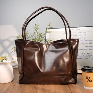 May include: A brown leather tote bag with long handles. The bag has a glossy finish and a rectangular shape. The background includes a white vase with flowers, a coffee cup with a percentage sign, and a handwritten note.