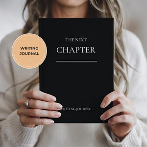 Peut inclure: Un journal d'écriture noir avec les mots "THE NEXT CHAPTER" en lettres blanches. Le journal est tenu par une personne portant un pull crème. Un autocollant rond sur la gauche indique "WRITING JOURNAL".