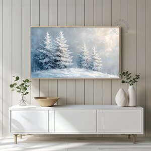 Könnte beinhalten: Ein gerahmtes Landschaftsgemälde zeigt schneebedeckte Tannenbäume in einer Winterszene. Das Kunstwerk weist eine hellblaue und weiße Farbpalette auf, mit einem Hauch von Sonnenlicht, das durch die Wolken bricht. Das Gemälde wird über einem weißen Schrank ausgestellt.