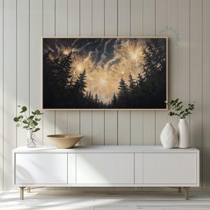Può includere: Una stampa incorniciata raffigura una scena notturna con fuochi d'artificio che esplodono sopra una sagoma di foresta. I fuochi d'artificio sono di colore giallo brillante e bianco contro un cielo blu scuro e nero. L'opera d'arte è esposta sopra un mobile bianco.