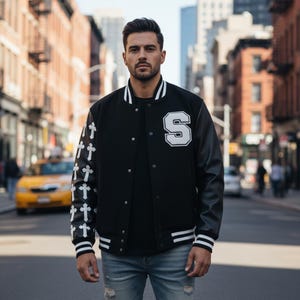 May include: Black varsity jacket with white accents and a large white "S" on the chest. The left sleeve features white crosses. The jacket has black leather sleeves and is worn with a black shirt and blue jeans.