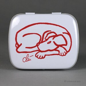 Puede incluir: Lata de metal blanca con un dibujo a línea roja de un perro durmiendo. La lata tiene una tapa con bisagras y está firmada "Drew" en la esquina inferior derecha.