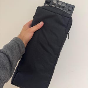 May include: A black keyboard inside a black padded carrying case. The keyboard has gray keycaps. The case has a zipper closure and is held by a person.