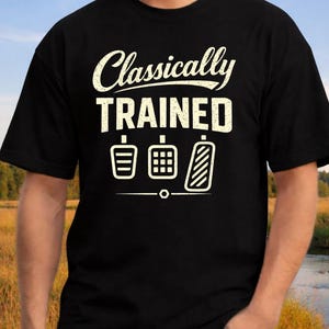 May include: Black t-shirt with the text "Classically Trained" in a distressed white font above three white graphic illustrations of car pedals. The shirt is a crew neck style.