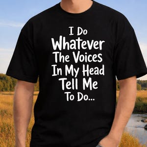Może przedstawiać: Czarny t-shirt z krótkim rękawem z białym napisem z przodu o treści: I Do Whatever The Voices In My Head Tell Me To Do... Koszulka ma okrągły dekolt i standardowy krój, co zapewnia prosty i swobodny wygląd w codziennym użytkowaniu.