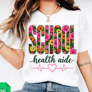 Puede incluir: Camiseta blanca con las palabras "SCHOOL" y "health aide" en un diseño floral. La palabra "SCHOOL" está llena de flores rosas, naranjas y amarillas. Una línea de latido de corazón rosa con un corazón está debajo.
