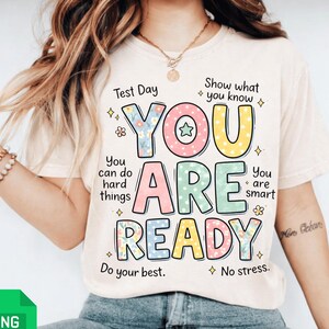 Puede incluir: Una camiseta de color beige claro con las palabras "YOU ARE READY" en letras de bloque coloridas. La camiseta tiene texto adicional que dice "Test Day", "Show what you know", "You can do hard things", "You are smart", "Do your best" y "No stress."