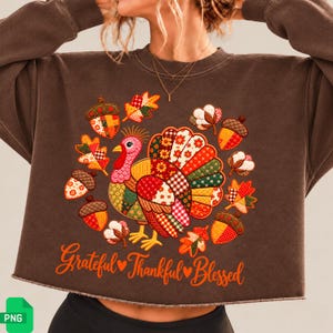Könnte beinhalten: Ein dunkelbraunes bauchfreies Sweatshirt mit einer zentralen Grafik eines Truthahns im Patchwork-Stil in Rot, Orange, Grün und Gelb. Der Truthahn ist von Eicheln, Herbstblättern und Baumwollblüten umgeben, darunter steht der Text Grateful Thankful Blessed in orangefarbener Schreibschrift.
