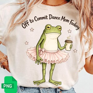 Può includere: T-shirt color crema con un ranocchio dei cartoni animati con un tutù rosa, che tiene in mano una tazza di caffè. Il testo "Off to Commit Dance Mom Foolery" è arcuato sopra il ranocchio, con stelle intorno.