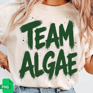 Peut inclure: Un t-shirt à manches courtes de couleur crème avec le texte "TEAM ALGAE" imprimé dans une police vert foncé au style pinceau vieilli. Le motif comprend de petites éclaboussures de peinture verte autour des lettres texturées et audacieuses.