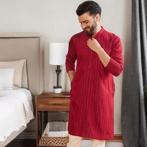 Op de afbeelding: Een rode kurta met verticale zilveren strepen, gecombineerd met crèmekleurige broek. De kurta met lange mouwen heeft een opstaande kraag en een knoopsluiting. De outfit wordt gecompleteerd met bruine sandalen. De kleding wordt in een slaapkamer getoond.