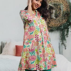 May include: A woman wearing a floral tunic and green pants. The tunic has a cream background with pink and green floral patterns. The pants are a solid green color. She is wearing light blue flats with gold trim.