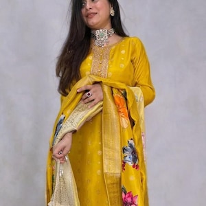Può includere: Una donna indossa un completo salwar kameez giallo senape. L'outfit presenta una tunica a maniche lunghe e pantaloni abbinati. Una sciarpa a fantasia floreale è drappeggiata sulle spalle. L'ensemble è impreziosito da una collana d'argento e una borsetta di perline.