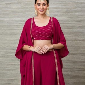 Könnte beinhalten: Ein magentafarbenes Dreiteiler-Outfit mit einem bauchfreien Top, Palazzo-Hose und einer langen, offen geschnittenen Jacke. Das bauchfreie Top ist mit kleinen, goldfarbenen Details verziert. Jacke und Hose haben goldfarbene Verzierungen. Das Outfit wird mit Goldschmuck getragen.