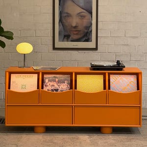 May include: A bright orange record cabinet with eight compartments, filled with vinyl records. A small, yellow, round lamp and a record player sit on top. A framed portrait hangs on the white brick wall behind the cabinet. A potted plant is on the left.