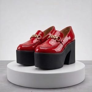 Pode incluir: Um par de mocassins de verniz vermelho com uma sola de plataforma preta alta e um detalhe de corrente dourada sobre o peito do pé. Os sapatos têm biqueira arredondada, salto bloco grosso, acabamento brilhante e uma silhueta estruturada.