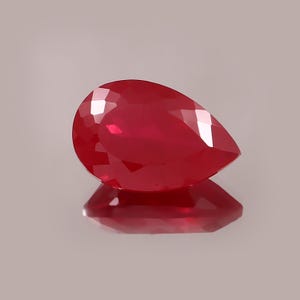 Puede incluir: Una gema de rubí facetada en forma de pera. La gema roja oscura está muy pulida, reflejando la luz y creando una exhibición brillante. La gema se asienta sobre una superficie reflectante, reflejando su forma y color.