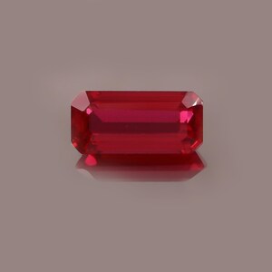 May include: A rectangular-cut, faceted ruby gemstone. The gem is a deep, saturated red colour with a high degree of clarity. The ruby is set against a neutral background, allowing its colour and brilliance to stand out. The gemstone is likely used in jewellery.